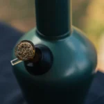 En slank bong i rustfrit stål med en højde på 33 cm. Overfladen har en poleret finish, hvilket giver en spejlende effekt. Den cylindriske form er enkel, med en afrundet top og en bredere bund. Der er en indbygget perkolator og et udluftningshul, samt en skåret kant ved åbningen. Farven er sølvagtig med en glat tekstur.