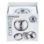 En kompakt grinder med en diameter på 65 mm, fremstillet af metal. Overfladen har en glat, blank finish med en sølvfarvet tone. Øverste del er udstyret med et skridsikkert mønster og en vinkelret kant. Der er synlige, skarpe tænder indeni til effektiv nedbrydning, samt et gennemsigtigt låg med en let buet form.
