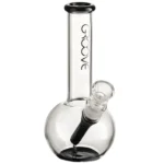 En rund bong i klart glas med en højde på 18 cm. Den har en slank, cylindrisk form med en bredere bund, der sikrer stabilitet. Der er en afbøjning i røret, der fører til et mundstykke, og en skål til urter er placeret på siden. Overfladen er glat, og der ses en subtil refleksion af lys.