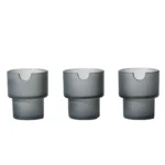 Askeholderen har en cylindrisk form med en glat, mat overflade i en dyb, mørkegrå farve. Den er fremstillet af keramik, hvilket giver en robust og elegant fremtoning. Øverste kant er let afrundet, og bunden er flad for stabilitet. Den har en diskret, indgraveret tekstur, der tilføjer et subtilt visuelt element til designet.