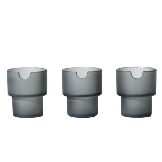 Askeholderen har en cylindrisk form med en glat, mat overflade i en dyb, mørkegrå farve. Den er fremstillet af keramik, hvilket giver en robust og elegant fremtoning. Øverste kant er let afrundet, og bunden er flad for stabilitet. Den har en diskret, indgraveret tekstur, der tilføjer et subtilt visuelt element til designet.