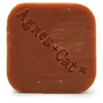 Den solide shampoo-bar har en rund, flad form med en glat overflade. Farven er en varm, lys orange, der udstråler en blød og cremet tekstur. Der er tydelige, subtile mønstre på overfladen, som tilføjer dimension. Baren prydes af en enkelt, stiliseret prikket detalje, der skaber visuel interesse uden at være overfyldt.