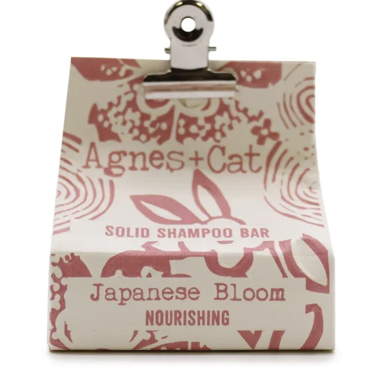 En solid shampoo bar med en rund form og en blød, let skinnende overflade. Farven er en delikat lyserød med subtile hvide striber. Overfladen har en let tekstur, som giver en fornemmelse af naturlige ingredienser. Der er ingen synlige emballageelementer, hvilket understreger det minimalistiske design. Produktet præsenteres på en lys, neutral baggrund.