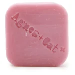 En solid shampoo-bar i en rund form præsenterer sig med en blød, glat overflade. Farven er en lys, sart nyanse af lyserød med små, fine blomsterdesigns indgraveret. Teksturen er let skinnende, hvilket indikerer fugt. Baren er placeret på en lys, naturlig overflade, der fremhæver dens bløde, minimalistiske udtryk.