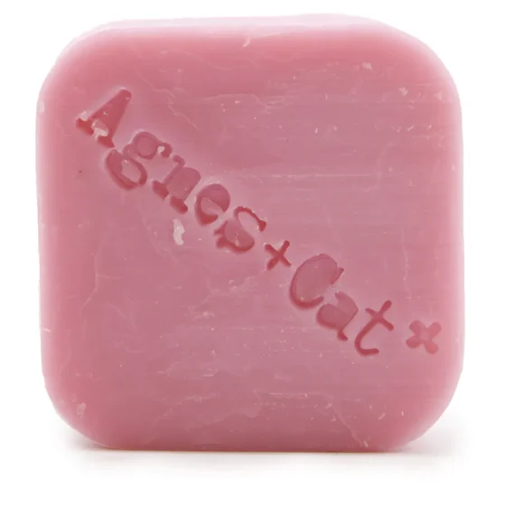 En solid shampoo-bar i en rund form præsenterer sig med en blød, glat overflade. Farven er en lys, sart nyanse af lyserød med små, fine blomsterdesigns indgraveret. Teksturen er let skinnende, hvilket indikerer fugt. Baren er placeret på en lys, naturlig overflade, der fremhæver dens bløde, minimalistiske udtryk.