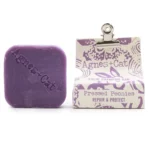 En solid shampoo-bar fremstår i en rund form, domineret af en blød beige farve med subtile nuancer. Overfladen er let struktureret, hvilket giver en mat finish. Der er indgraverede detaljer, som minder om blomstermønstre, der tilføjer en dekorativ dimension. Baren ligger på en naturlig, lys træoverflade, der kontrasterer med produktets farvetoner.