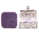 En solid shampoo-bar fremstår i en rund form, domineret af en blød beige farve med subtile nuancer. Overfladen er let struktureret, hvilket giver en mat finish. Der er indgraverede detaljer, som minder om blomstermønstre, der tilføjer en dekorativ dimension. Baren ligger på en naturlig, lys træoverflade, der kontrasterer med produktets farvetoner.