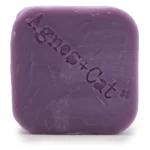 En fast shampoo-bar præsenteres med en oval form og en glat, ensartet overflade. Farven er en blød, lyserød nuance, der minder om pasteller. Overfladen har en let glans, og der ses små, fine indtryk, der skaber tekstur. Baren er pakket i en simpel, miljøvenlig indpakning med grønne detaljer og plantemotiver, der fremhæver naturlige ingredienser.