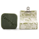 En solid shampoo-bar i en rund form, fremstillet af hvidt materiale. Overfladen har en glat tekstur med en let skinnende finish. Der er indgraveret et simpelt, elegant mønster på toppen, hvilket tilføjer visuel interesse. Shampoo-baren er placeret på en neutral, lys overflade, hvilket fremhæver dens farve og form.