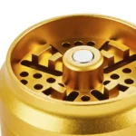 En gylden krydderkværn med en diameter på 55-63 mm fremstår i et slebet metalfinish, der reflekterer lys. Overfladen er glat med fine detaljer, som små riller langs kanten. Kværnen har en skruelåg med et indgraveret mønster, der tilføjer tekstur. Indvendigt ses skarpe tænder til formaling af urter. Kværnen er kompakt og robust i design.