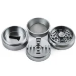 Grinderen præsenterer en sølvfarvet metaloverflade med en glat og skinnende finish. Den har en cirkulær form med en diameter på 55-63 mm og er udstyret med en skarp tandet inderside til effektiv kværning. Toppen har et skridsikkert mønster, og der er små ventilationshuller langs kanten. En lille, diskret logo er placeret på overfladen.