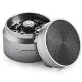 Grinderen har en sølvfarvet metaloverflade med en glat, skinnende finish. Den er cylindrisk med en diameter på 55-63 mm. Øverste del har et skruelåg med små, skarpe tænder indvendigt, der er designet til at male. Nederste del er flad med en lille åbning til opbevaring. Der ses fine ridser og et let refleksionseffekt.