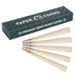 En pakke med 32 hamp cones præsenteres i en rektangulær, genanvendelig papæske. Æsken har en grønlig farve med et minimalistisk design. På forsiden ses en enkel, hvid tekst, der angiver produktets navn. Indeni ligger cones, der er rullet i et naturligt papir, tydeligt synlige med en let, ru tekstur. De er ensartede og har en spids top.