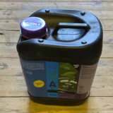 En klar plastflaske med et væskeindhold, der har en dyb, mørkegrøn farve. Flasken har en slank, cylindrisk form og en skruelåg i hvid plast. Etiketens design er enkelt, med tekst i sort og grøn, der angiver produktinformation. Flasken står på en neutral overflade, hvilket fremhæver dens farver og former.