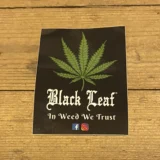 Klistermærket præsenterer en sort baggrund med en glat, mat overflade. Centralt står teksten "In Weed We Trust" i en kontrastfyldt hvid farve, skrevet med en fed, moderne skrifttype. Øverst er der et stiliseret blad, der symboliserer cannabis, i grønne nuancer. Mønstrene og skrifttypen giver et ungdommeligt og uformelt udtryk, der tiltaler en bestemt målgruppe.