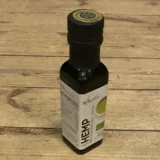 En transparent flaske med en mørk grøn væske indeholder hampolie. Flasken har en enkel, cylindrisk form med en sort skruelåg, der har en lille åbning til dosering. Etiket med hvid baggrund pryder flaskens front, indeholdende grøn tekst og symboler, der angiver produktinformation. Flasken står på en neutral overflade, hvilket fremhæver dens farver.