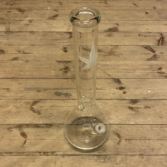 En høj, transparent bong i klart glas med en slank, cylindrisk form. Den har en hvid, bladformet dekoration, der strækker sig op ad siden. Toppen er bred med en let udvidelse, mens bunden er stabil og rundet. Der er en åbning til røg og en fordybning til vand, som sikrer stabilitet og funktionalitet.