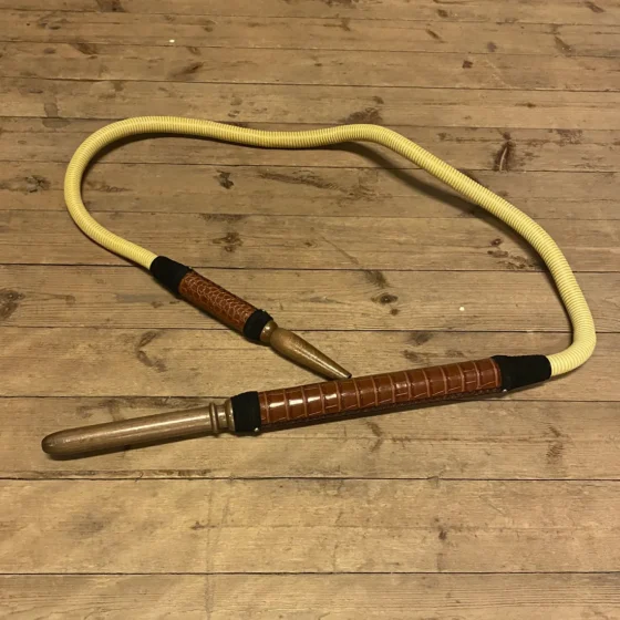 En traditionel tyrkisk slange med et mørkebrunt læderhåndtag præger designet. Slangen har en elegant, slank form og er dækket af et mønster i nuancer af grå og beige, hvilket giver en tekstureret overflade. Håndtaget er lavet af blødt læder med synlige syninger, der tilføjer detaljer og kontrast til slangen.