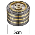 En sølvfarvet grinder med en blank overflade, der reflekterer lys. Den har en diameter på 50 mm og er udstyret med flere skarpe tænder indeni til effektiv formaling. Låget prydes af et detaljeret, farverigt bitcoin-symbol, der tilføjer et grafisk element. Grinderens kanter er let afrundede for ergonomisk greb.