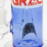 En 28 cm høj bong i klart glas præsenterer en elegant, buet form med en bred bund, der gradvist snævrer ind mod toppen. Indvendige detaljer omfatter en indbygget perkolator, der skaber bobler. Mønstret af en haj pryder glasset med en kombination af blå og hvide nuancer, hvilket tilføjer visuel dybde. Kanten er forsynet med en glat, ensartet finish.