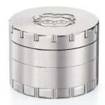 En grå, cylindrisk kværn i rustfrit stål med en diameter på 63 mm. Den har en glat overflade og en let reflekterende finish. Kværnen er opdelt i to sektioner, med en skruelås og små, skarpe tænder indeni, der er designet til at male urter. En lille åbning til opbevaring er placeret i bunden.