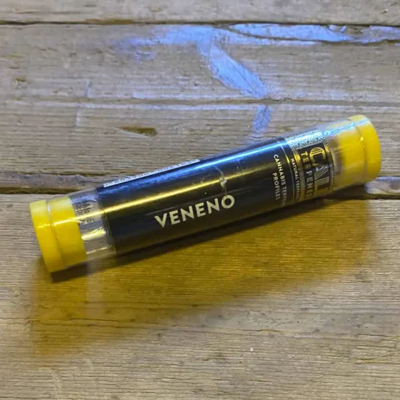 En lille glasflaske med en slank, cylindrisk form, fyldt med en klar væske. Flasken har en sort skruelåg med indgravering. Et etikette i lyse farver pryder flaskens front, hvor der står "Veneno" i store, fremtrædende bogstaver. Baggrunden er neutral, hvilket fremhæver flaskens detaljer og farver.