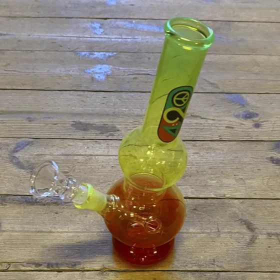 En 25 cm høj bong med en rasta-inspireret farveskala, der kombinerer grønne, gule og røde nuancer. Den har en glat, gennemsigtig krop med en krum hals og en bred bund. Bundens indre har en perforeret skive, som er synlig. Der er en indsat for røgen, og toppen har en bred åbning. Overfladen er skinnende og glat.