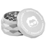 En sølvfarvet hex grinder med en diameter på 55 mm præsenterer sig med en glat, metalisk overflade. Den har en sektioneret konstruktion med flere skarpe tænder indeni, der er designet til at knuse materiale. Låget har en let buet form, og et lille hul til udledning af det hakkede indhold er placeret i bunden.