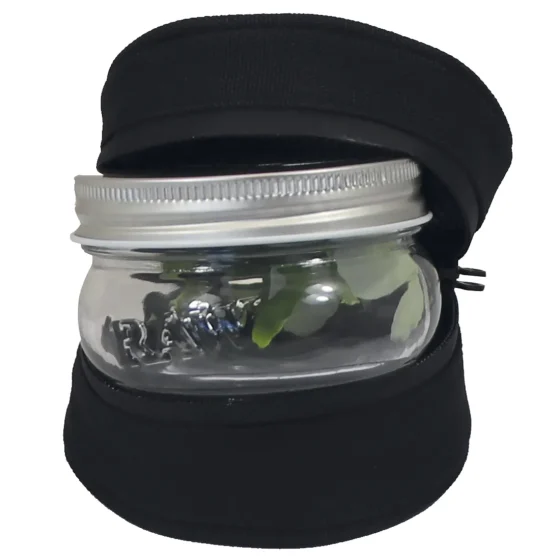 Et klart glas mason jar med en glat overflade og en rund, bred åbning. Låget er metalisk med en sølvgrå finish, der har en let struktur. Jarret er placeret i en beskyttende, mørkebrun stofpose, som har en blød tekstur og snøring. Posen dækker bunden af glasset og skaber en kontrast til de glatte glas- og metaloverflader.