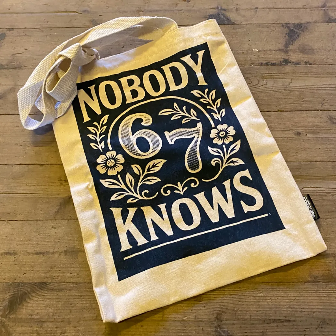 Nobody Knows Tote Bag m. Lynlås 32x42 cm i beige stof med sort trykfelt, blomster og teksten "NOBODY KNOWS".
